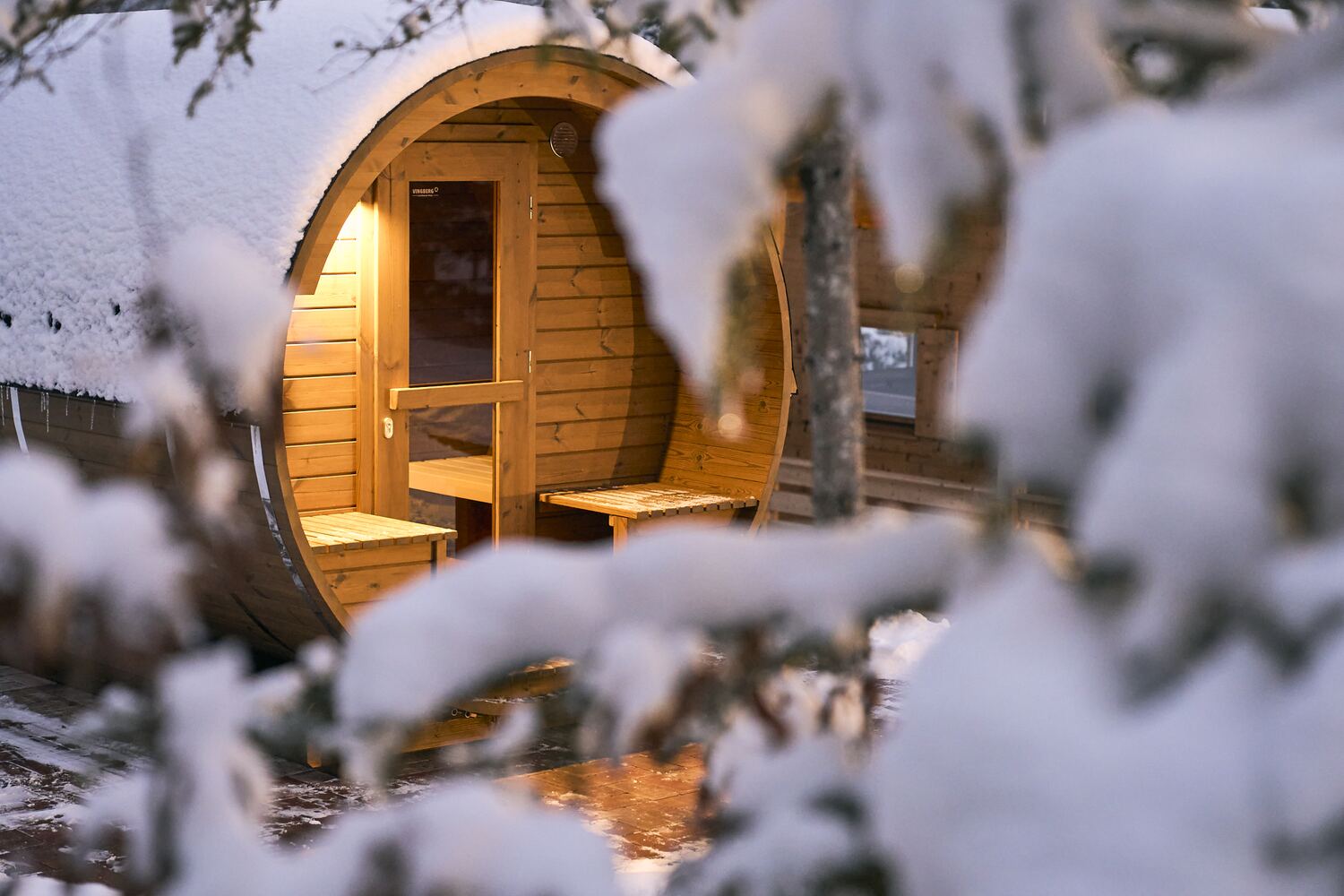 Warm beleuchtete Fass-Sauna im verschneiten Wald bei Dämmerung